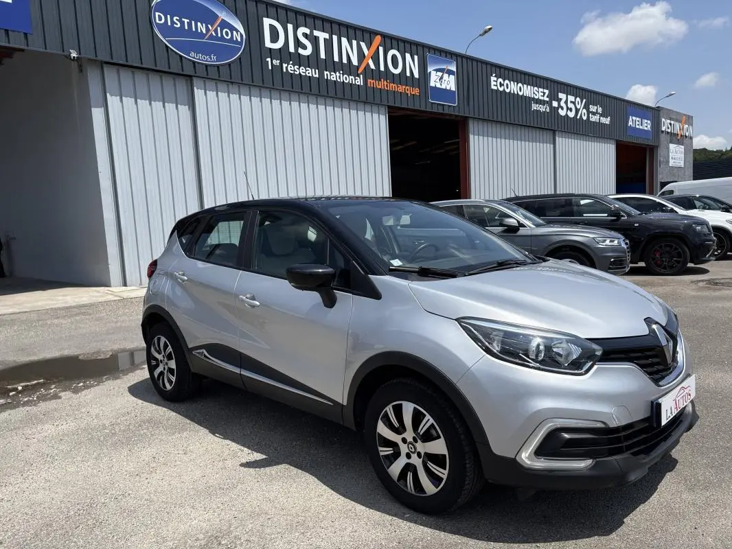 Renault Captur gris clair vue 3/4 avant droit, toit noir, jantes aluminium, stationné devant un garage multimarque.