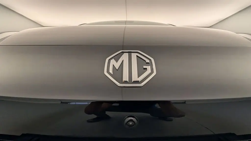 Gros plan sur le logo MG au centre d'une malle arrière Pebble Black Metal, avec caméra de recul visible en dessous.