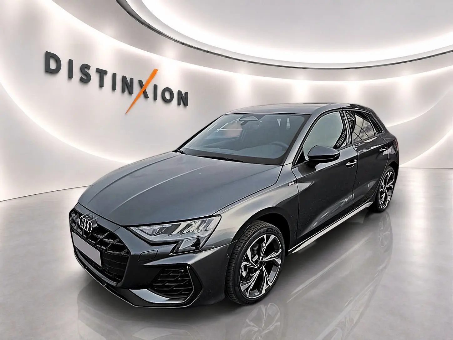 Audi A3 Sportback gris Daytona vue 3/4 avant droit dans un showroom lumineux avec logo Distinxion en arrière-plan.