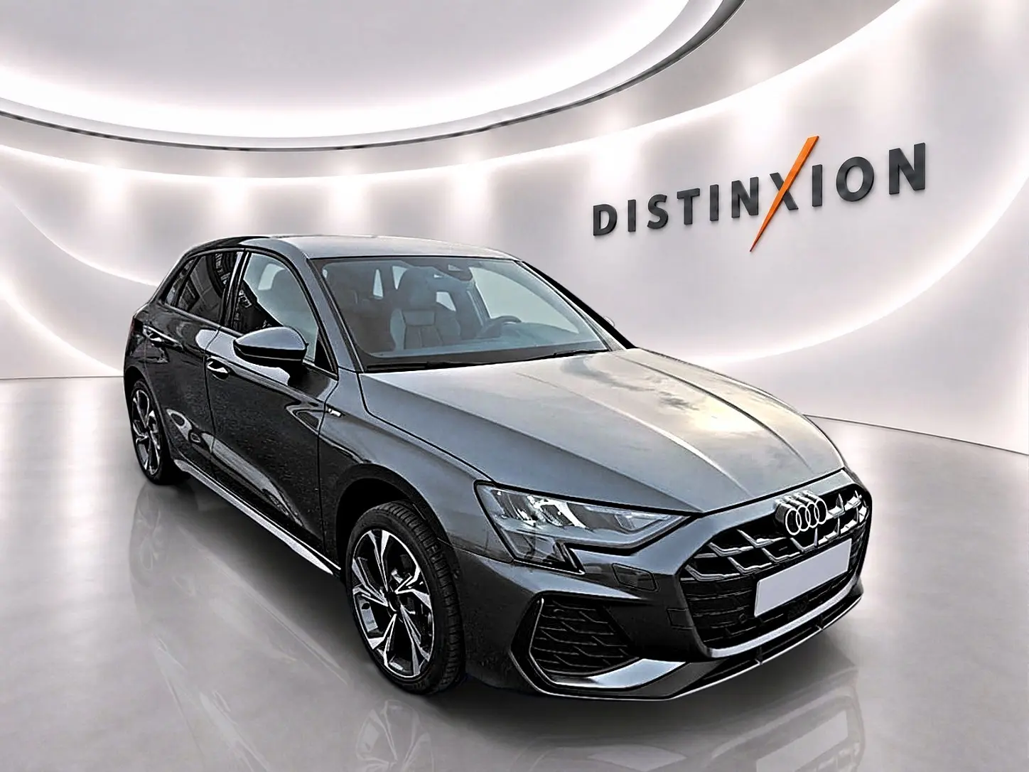 Audi A3 Sportback gris Daytona vue 3/4 avant droit dans un showroom lumineux avec logo Distinxion en arrière-plan.