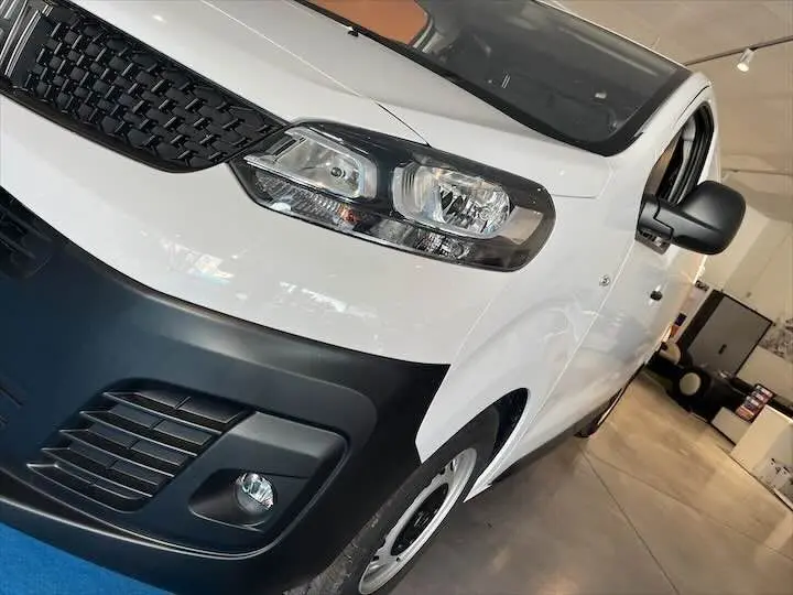 Vue 3/4 avant droit d'un FIAT Scudo Fourgon blanc avec pare-chocs noir et phares modernes en intérieur showroom.