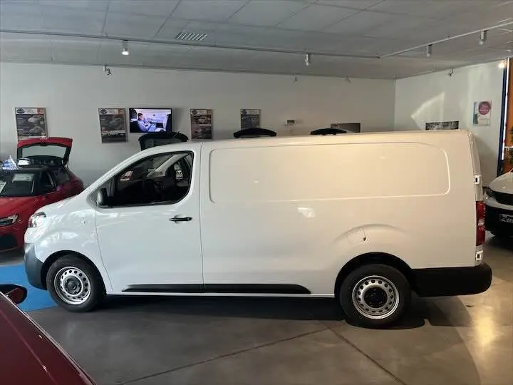 Profil côté gauche du FIAT Scudo Fourgon blanc, version longue, dans un showroom lumineux avec d'autres véhicules visibles.
