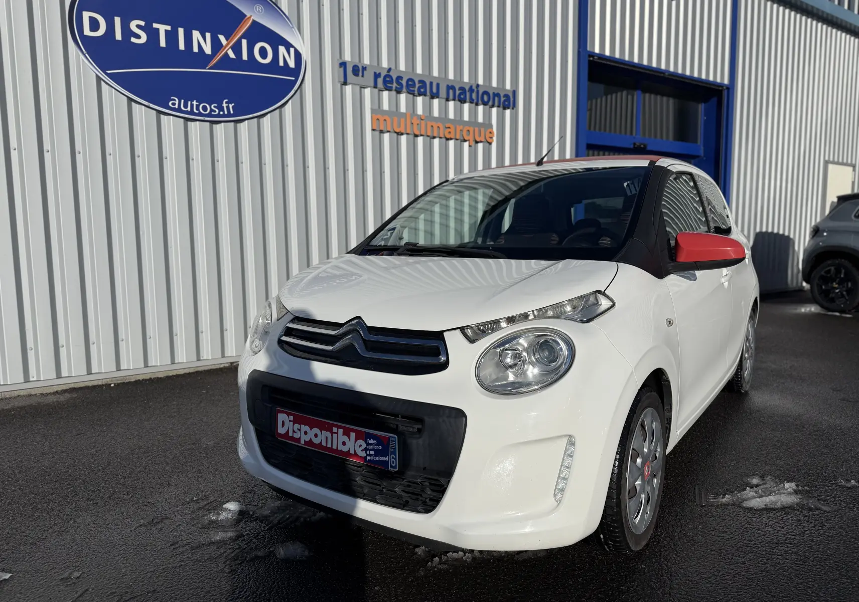 Citroën C1 blanc vue 3/4 avant droit avec rétroviseurs rouges, stationnée devant un bâtiment commercial.