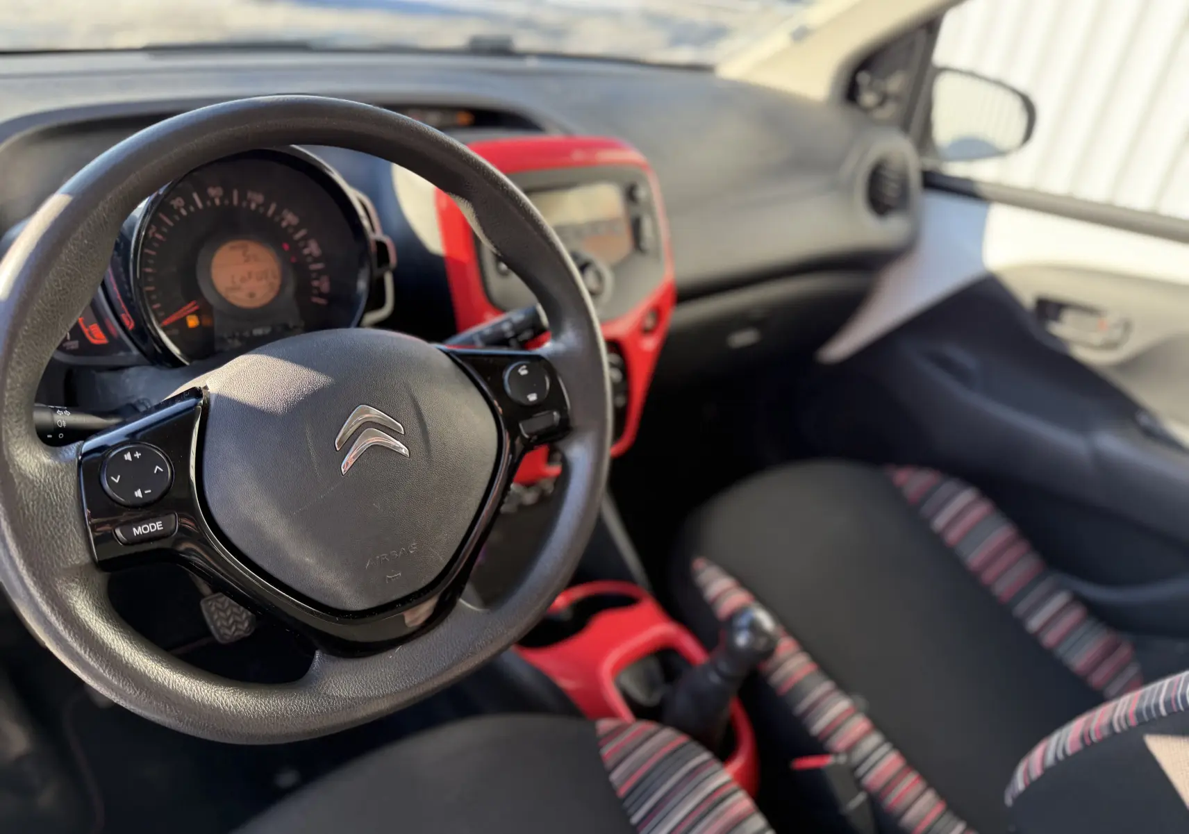 Vue intérieure côté conducteur d'une Citroën C1 2014 avec volant multifonction, tableau de bord noir et touches rouges, sièges tissu rayé.