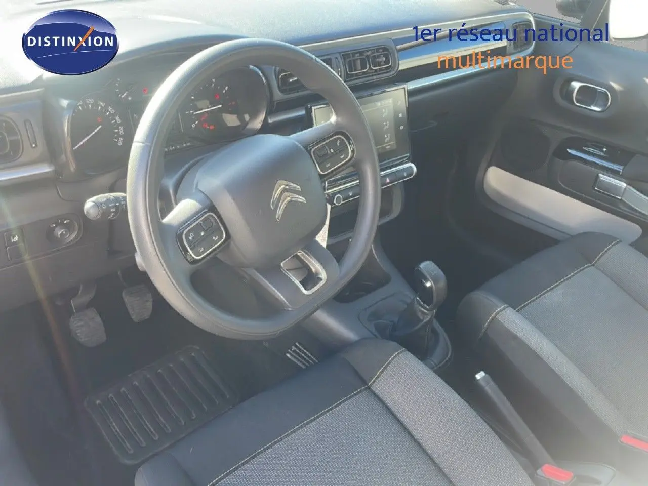 Vue intérieure de face du poste de conduite d'une Citroën C3 blanche, volant multifonction et levier de vitesse manuel visibles.