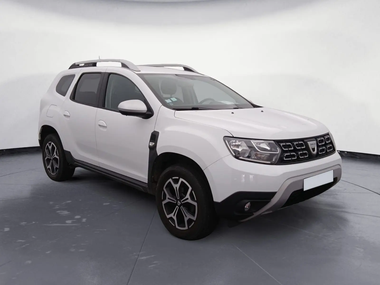 Dacia Duster blanc en 3/4 avant droit, avec calandre noire et jantes alliage, stationné sur un sol gravillonné.