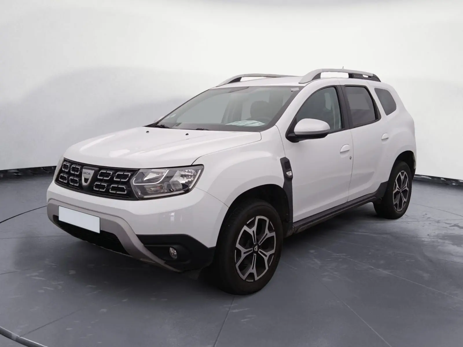 Dacia Duster blanc vue 3/4 avant droit sur parking, avec jantes alliage noires et toit barres de toit grises.