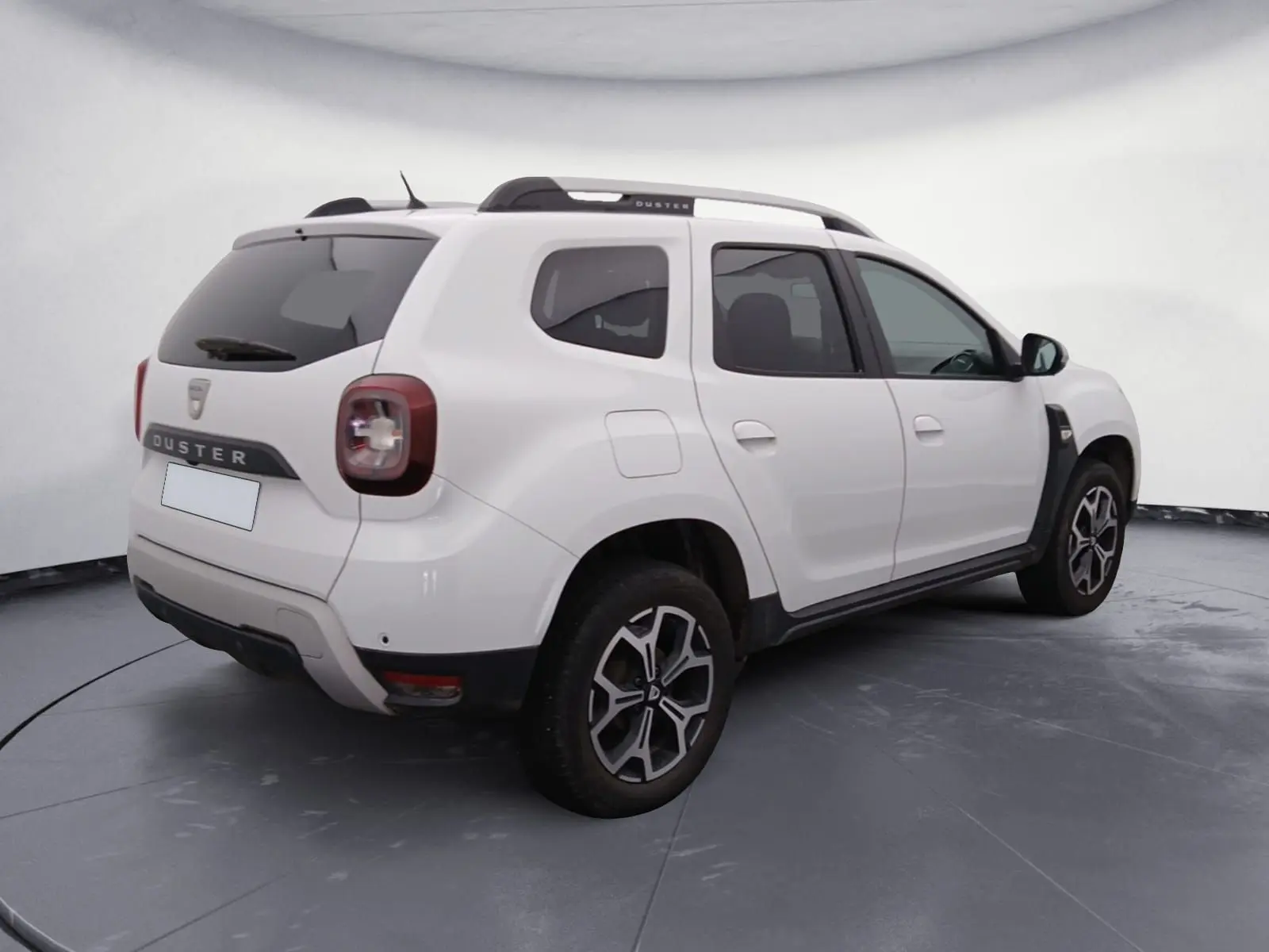 Vue 3/4 arrière droite d’un Dacia Duster blanc 2021 avec jantes alliage noires et toit ouvrant, stationné sur un terrain gravelé.