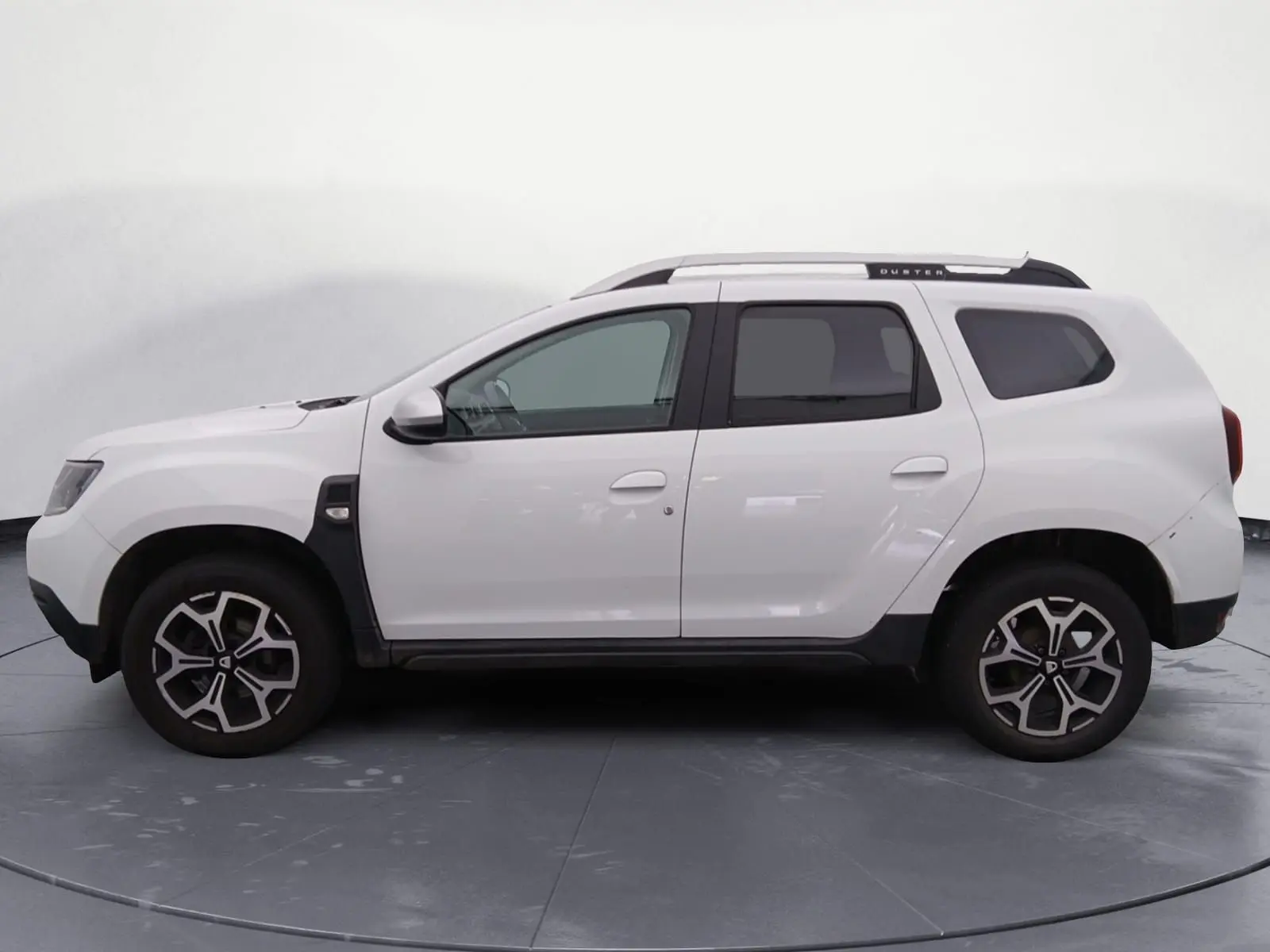 Vue 3/4 arrière droite du Dacia Duster blanc 2021 avec jantes alliage noires et barres de toit visibles sur un terrain gravelé.
