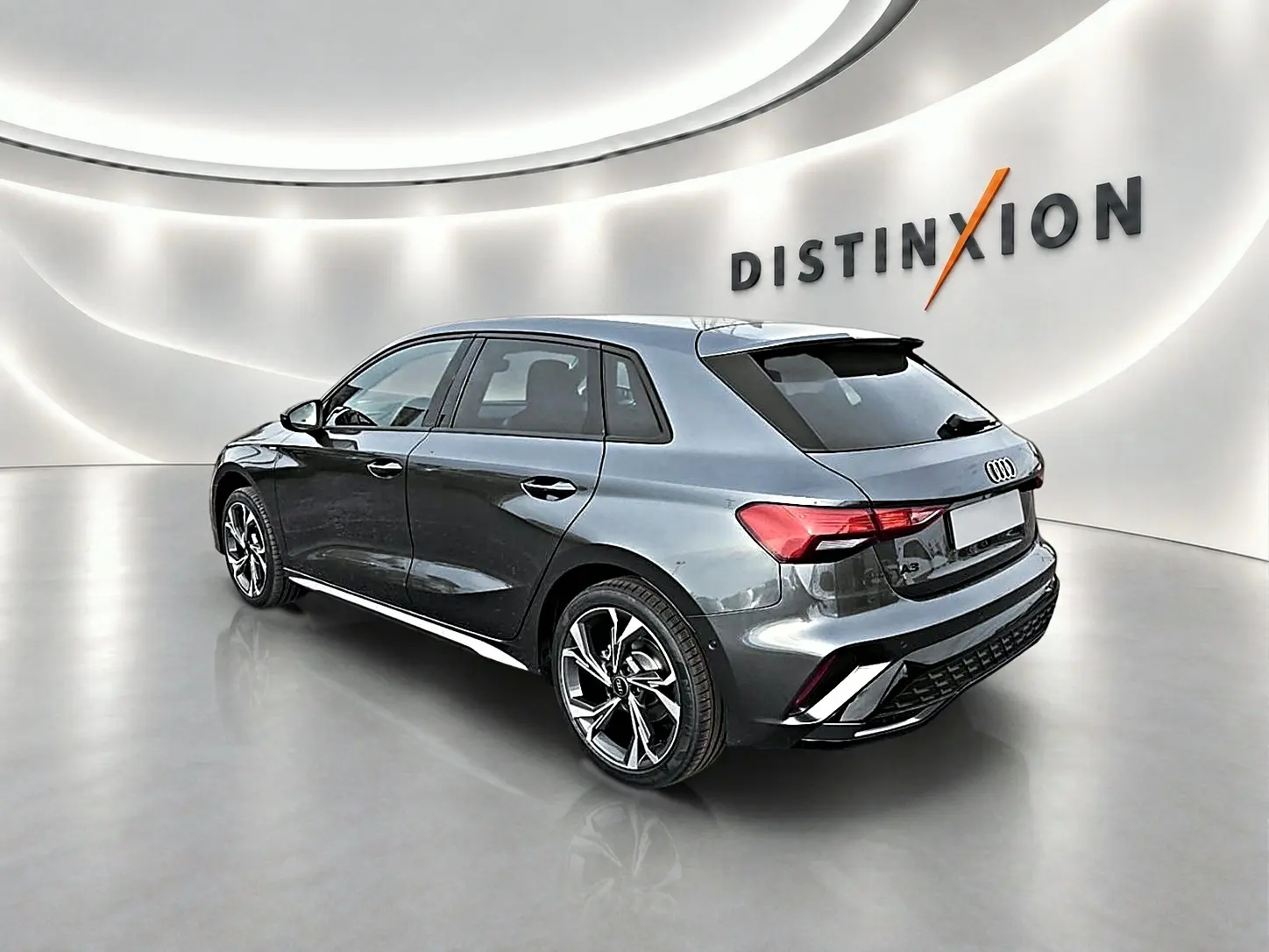 Audi A3 Sportback gris Daytona vue 3/4 arrière droit dans un showroom lumineux avec jantes alu noires et becquet arrière.
