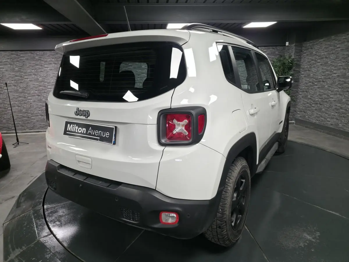 Vue 3/4 arrière droite d'un Jeep Renegade blanc avec jantes noires 5 branches et barres de toit gris clair.