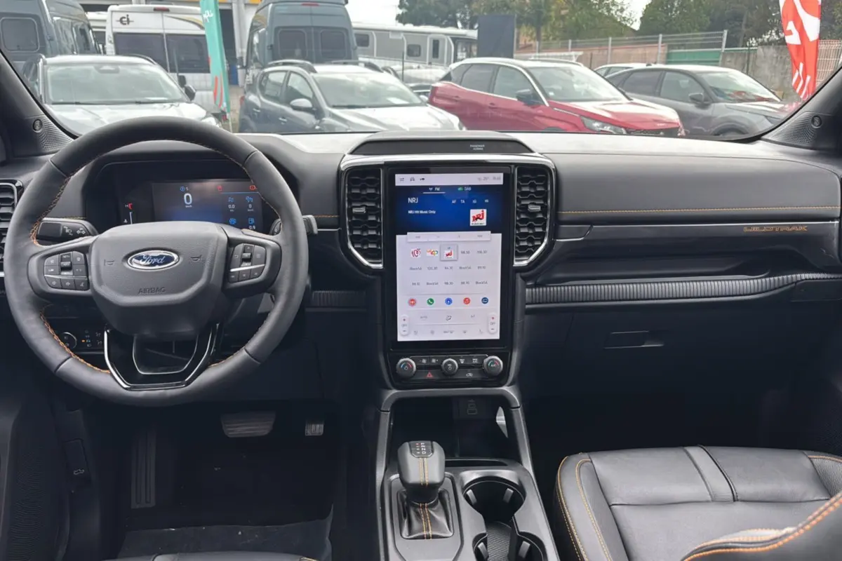 Vue intérieure avant du Ford Ranger Wildtrak Plus 2025 noir agate, avec volant cuir, écran tactile vertical et surpiqûres orange.