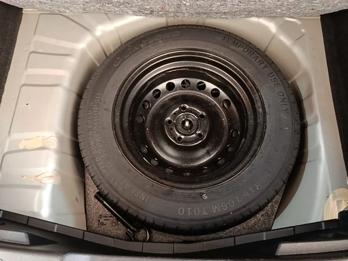 Roue de secours galette noire dans le coffre d’un Nissan Juke gris clair, vue plongeante centrée.