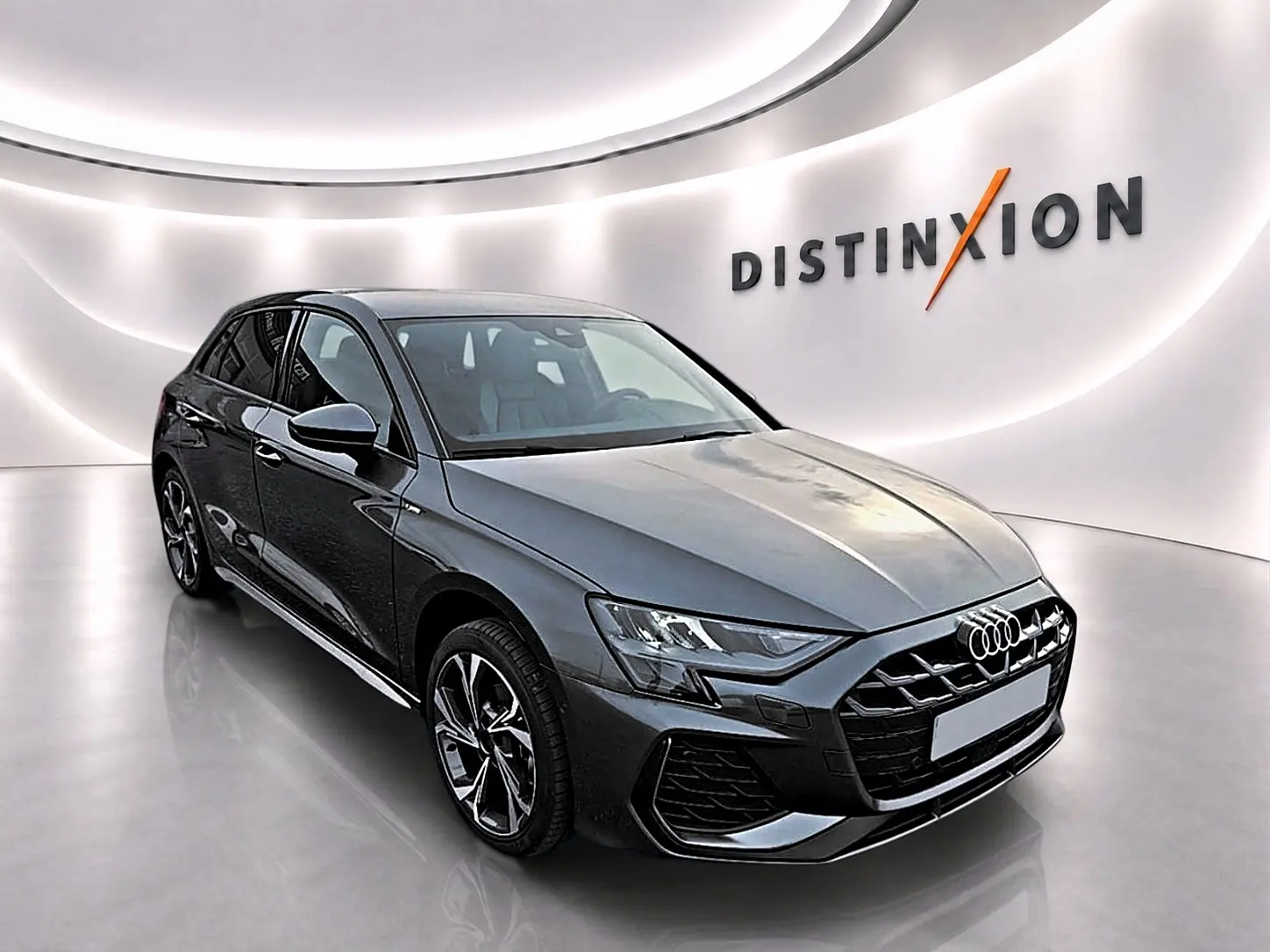 Audi A3 Sportback gris Daytona vue 3/4 avant droit dans un showroom lumineux avec logo Distinxion en arrière-plan.