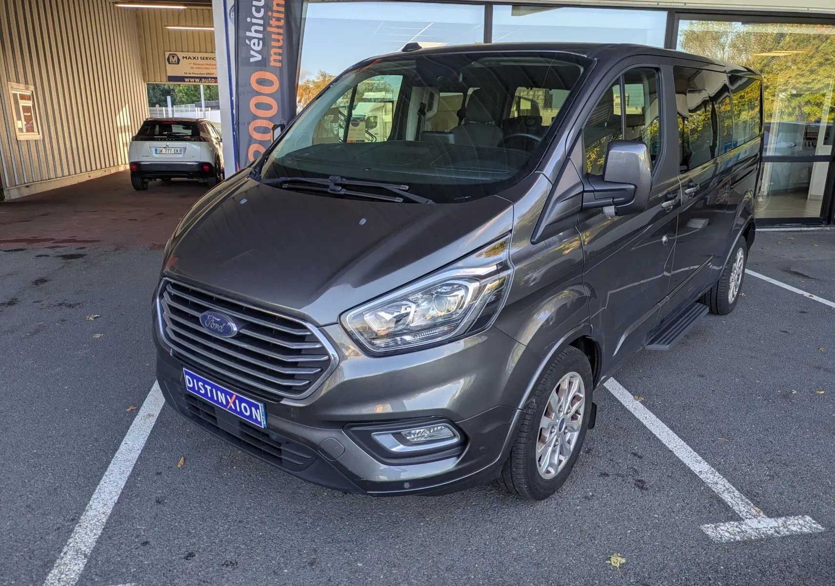 Ford Tourneo Custom gris en 3/4 avant droit, avec calandre chromée et jantes alliage, stationné devant un bâtiment.