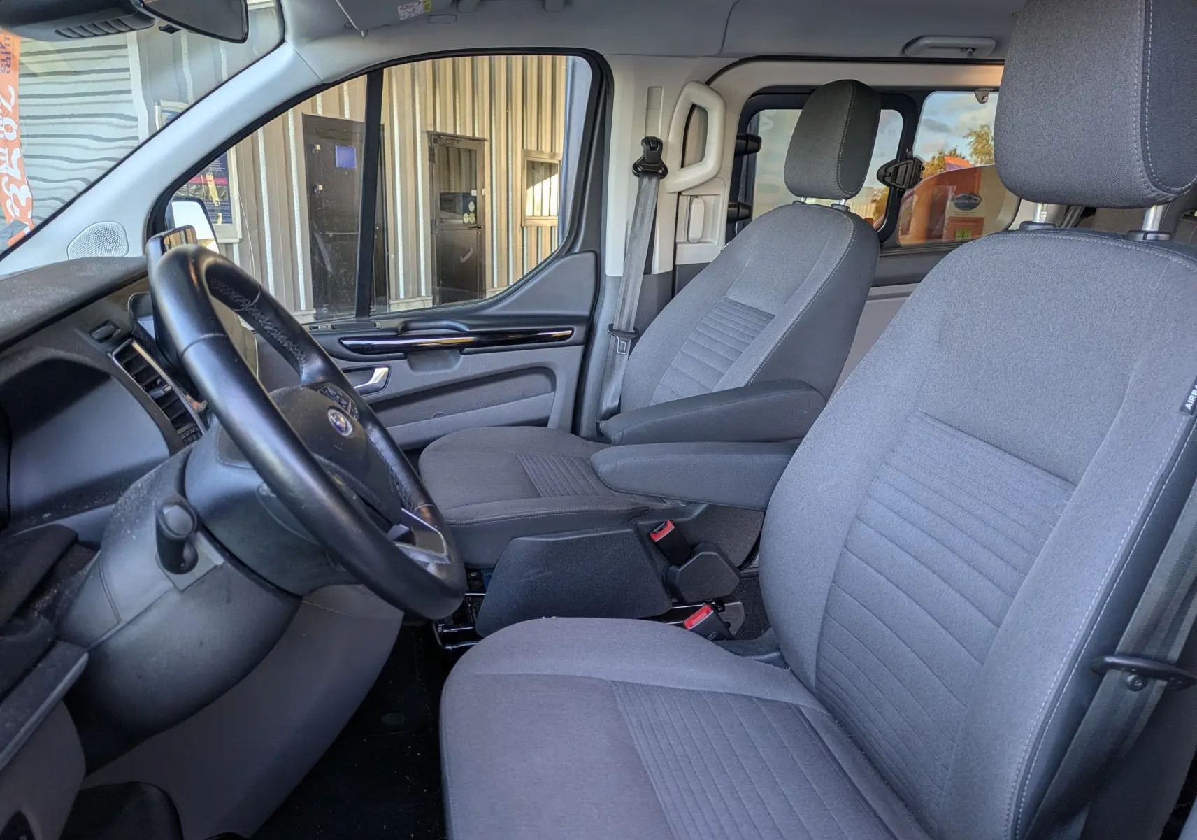 Vue intérieure côté conducteur du Ford Tourneo Custom gris 2019, montrant les sièges avant et le volant noir.
