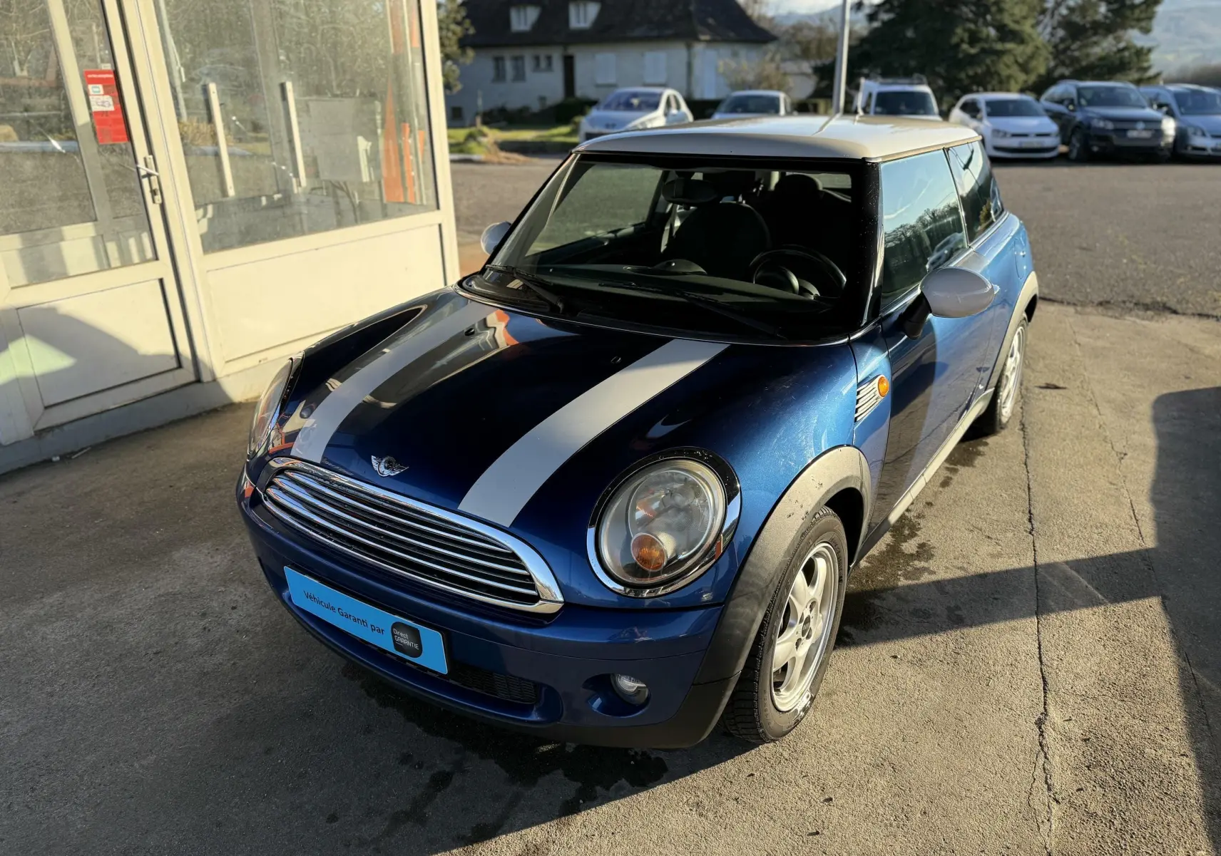 MINI Cooper bleu avec bandes blanches vue de 3/4 avant côté gauche, garée devant un bâtiment ensoleillé.