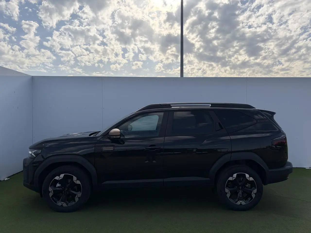 Profil côté gauche du Dacia Bigster noir nacré 2025 avec jantes alliage et rétroviseurs brun cuivré sous ciel nuageux.