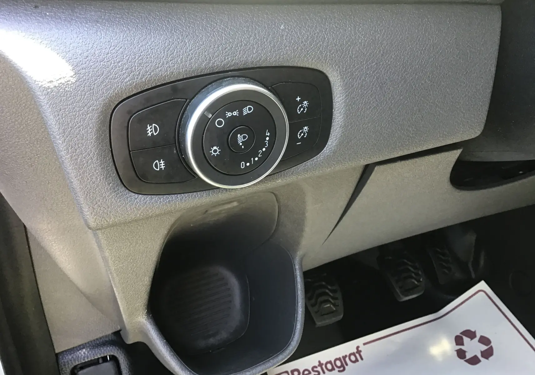 Gros plan sur le bouton de réglage des phares à gauche du tableau de bord du Ford Transit Custom blanc, avec pédales visibles en arrière-plan.