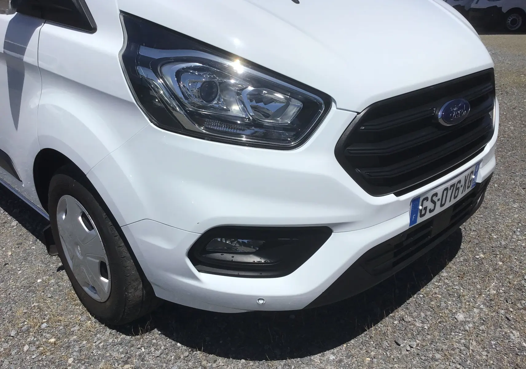 Vue 3/4 avant droit du Ford Transit Custom Kombi blanc, mettant en valeur le phare et la calandre noire.
