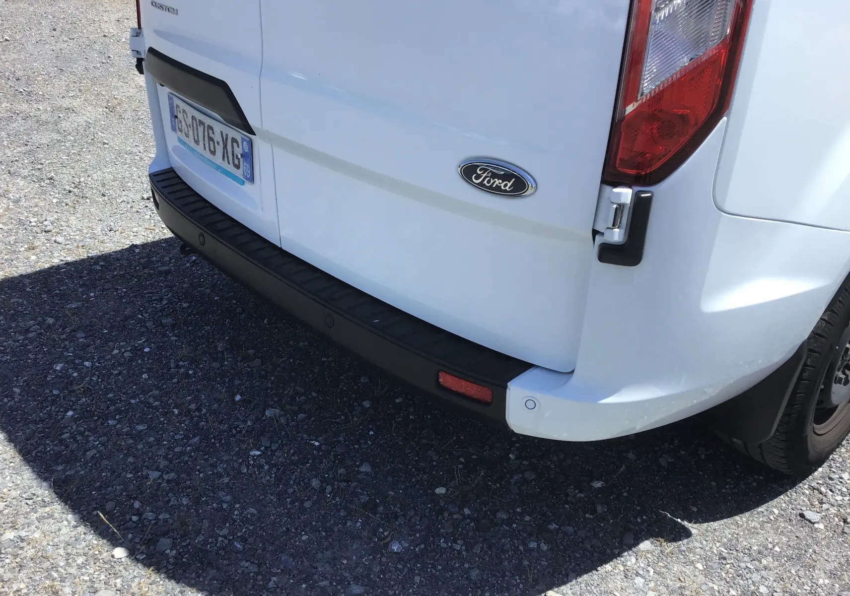 Vue rapprochée arrière droite du Ford Transit Custom Kombi blanc, mettant en valeur le logo et le pare-chocs noir.