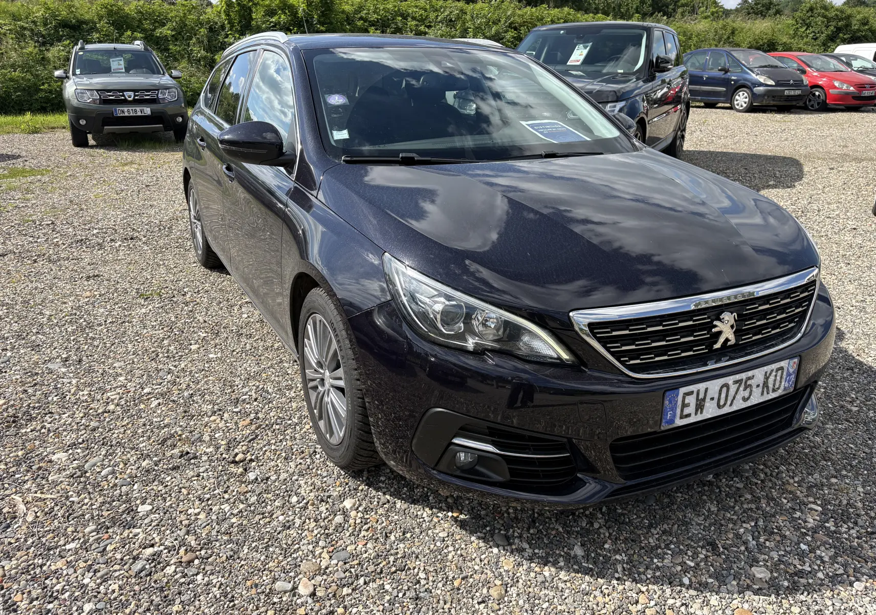Peugeot 308 SW noire vue en 3/4 avant droit sur un parking gravillonné avec plusieurs autres voitures en arrière-plan.