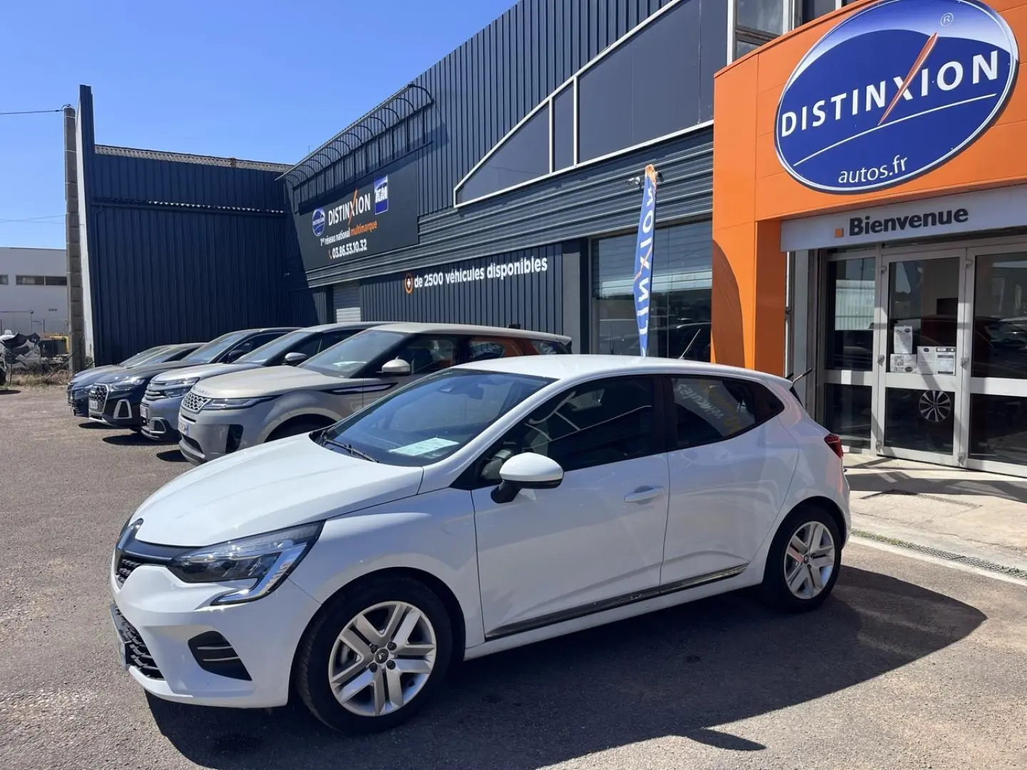 Vue 3/4 avant droite d'une Renault Clio blanc quartz garée devant un showroom avec enseigne orange Distinxion.