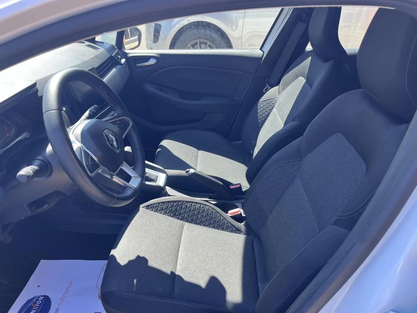 Vue intérieure côté conducteur de la Renault Clio blanc quartz, sièges tissu noir avec surpiqûres et volant multifonction.