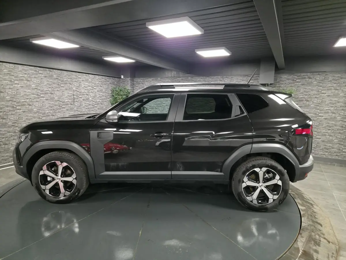 Profil droit du Dacia Duster 1.6 Hybrid 2025 noir nacré avec jantes alliage 18 pouces et barres de toit noires.