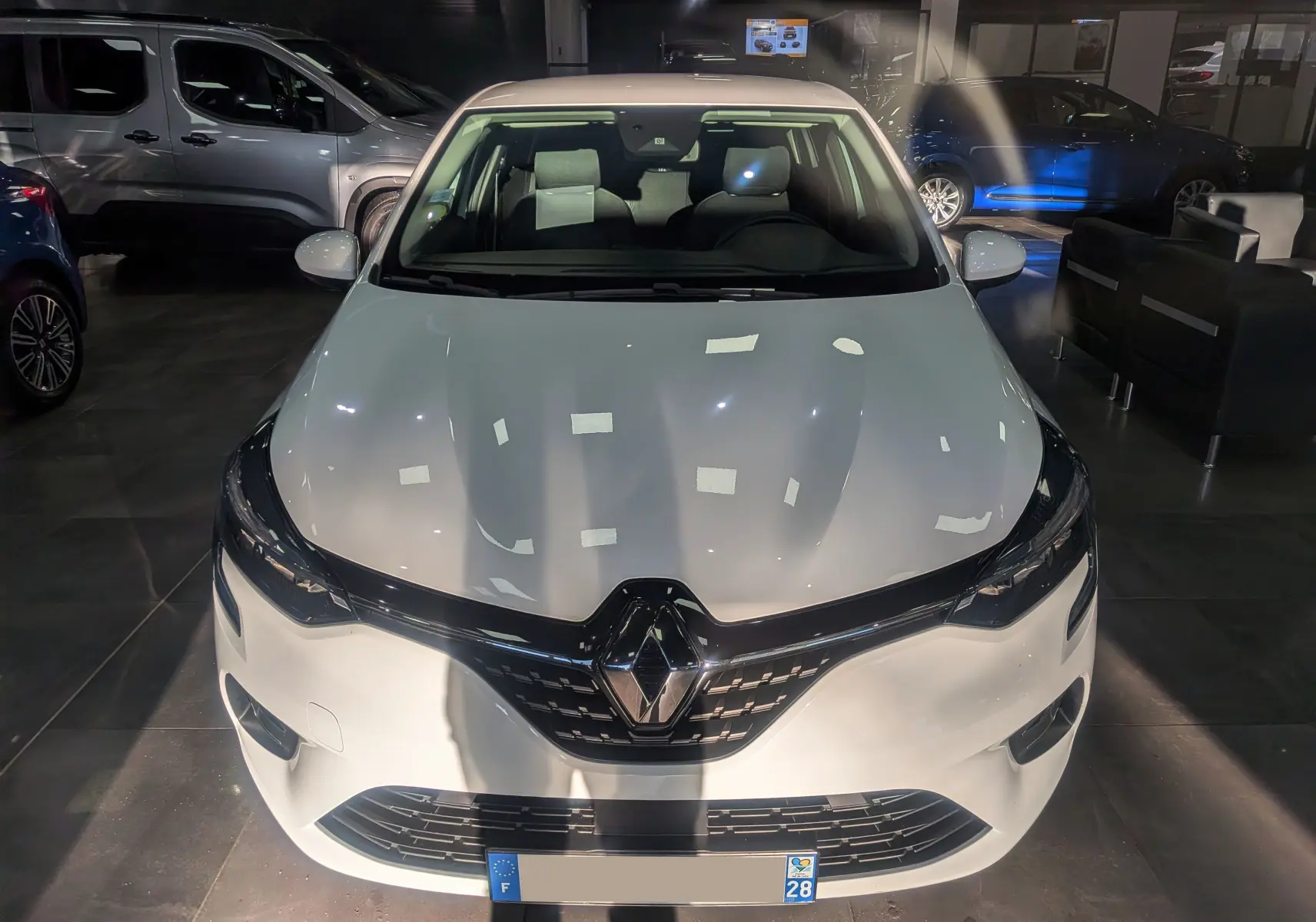 Vue avant d'une Renault Clio V Blanc Glacier 2021 en showroom, avec calandre noire et logo Renault central visible.