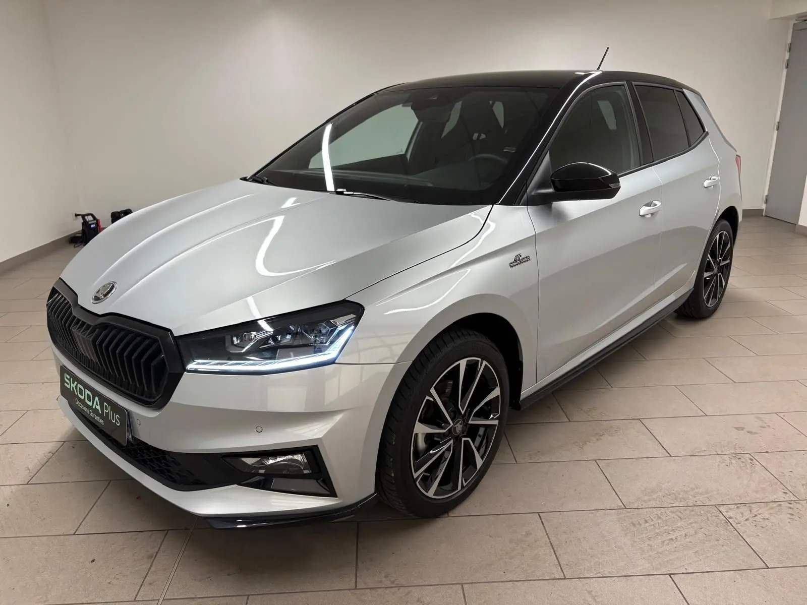 Vue 3/4 avant d'une Skoda Fabia 2025 gris argent avec toit noir et jantes noires, en intérieur sur sol carrelé.