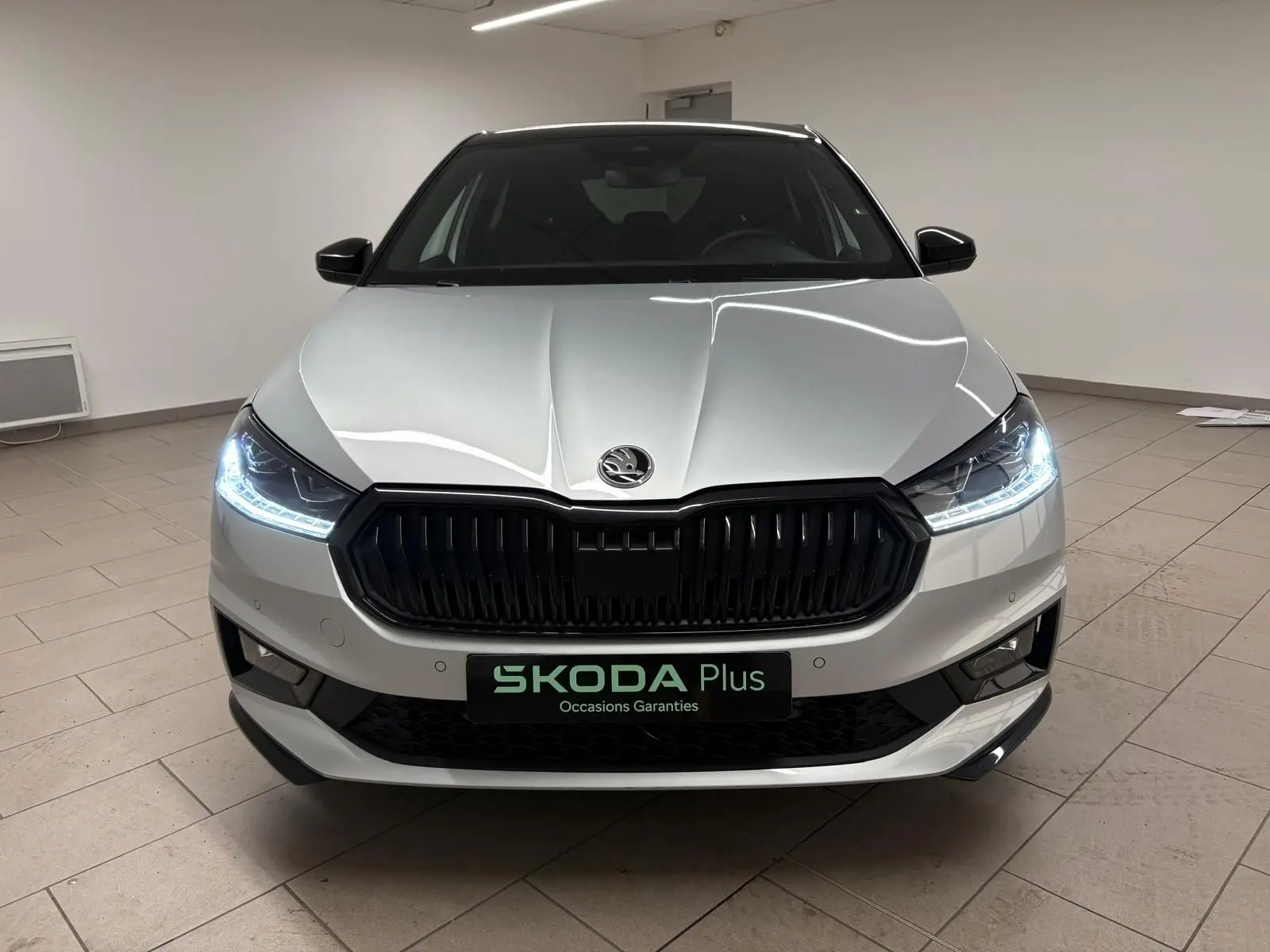 Vue frontale d'une Skoda Fabia 1.0 TSI 116 DSG7 Monte Carlo gris argent avec toit noir et phares allumés en intérieur.