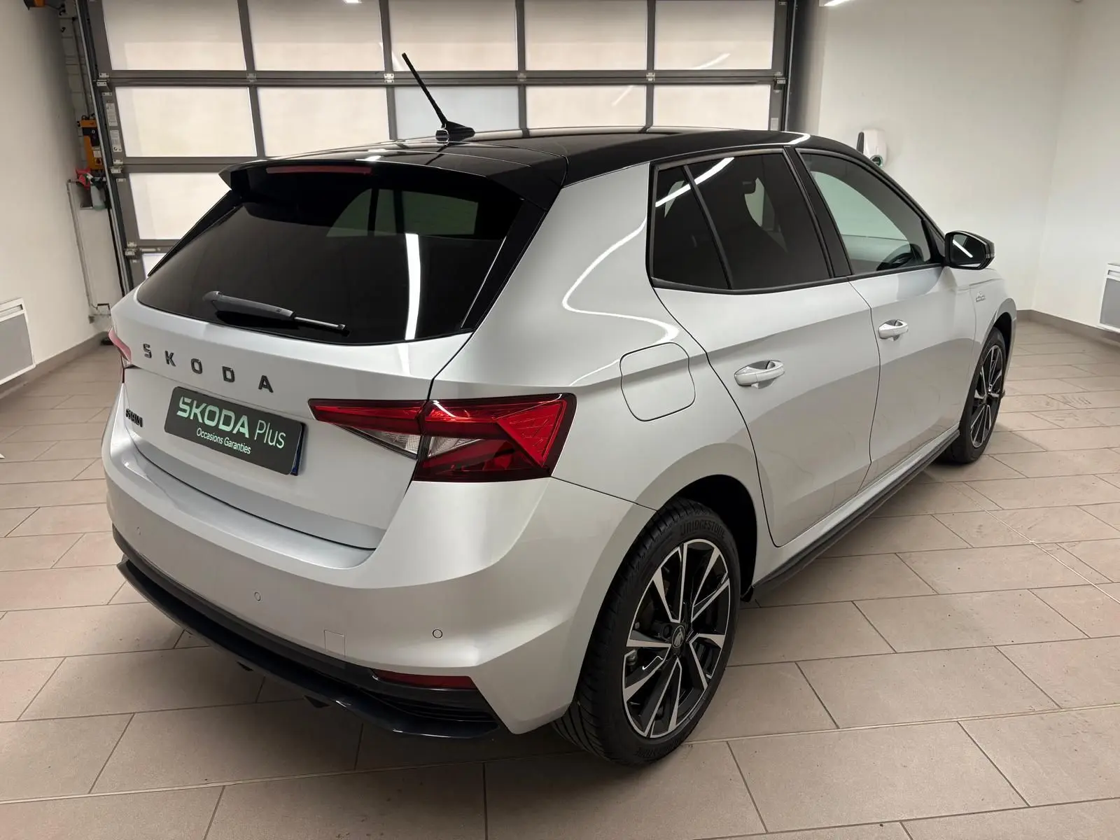 Vue 3/4 arrière droite d'une Skoda Fabia 2025 gris argent avec toit noir et jantes alliage noires en intérieur showroom.