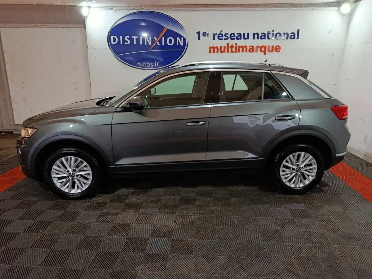 Vue latérale droite d'un Volkswagen T-Roc gris foncé 2021 dans un showroom avec logo Distinxion en arrière-plan.