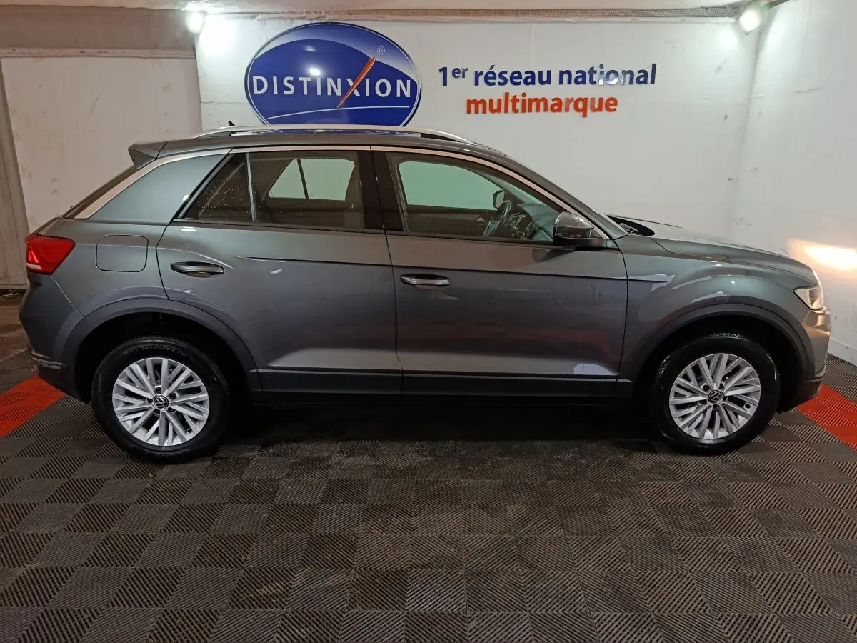 Vue de profil côté gauche d'un Volkswagen T-Roc gris foncé 2021 dans un showroom avec logo Distinxion en arrière-plan.