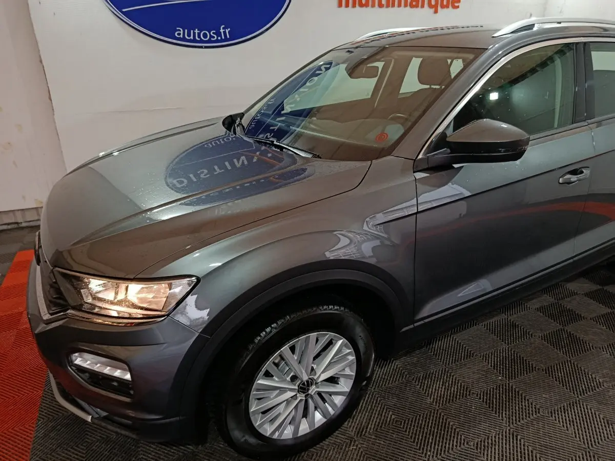 Vue 3/4 avant droit du Volkswagen T-Roc gris foncé avec phares allumés et jantes alliage visibles.