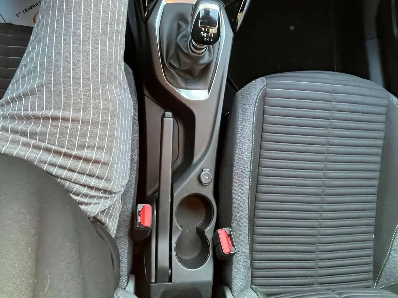 Vue plongeante sur la console centrale d'une Peugeot 2008 2024, boîte manuelle, sièges gris et boutons rouges des ceintures.
