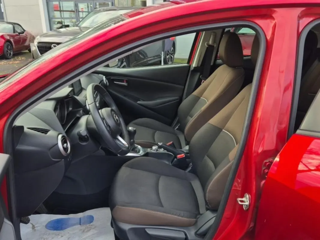Vue intérieure côté conducteur de la Mazda2 rouge 2021, sièges bi-ton marron et noir avec volant multifonctions cuir.