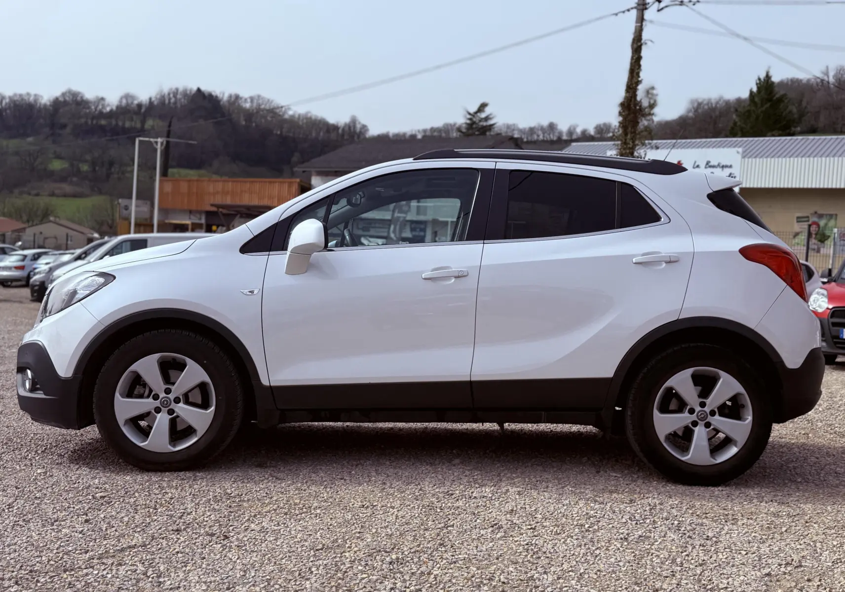 Vue de profil côté gauche d'un Opel Mokka X blanc avec jantes alliage et vitres teintées, stationné sur un sol gravillonné.