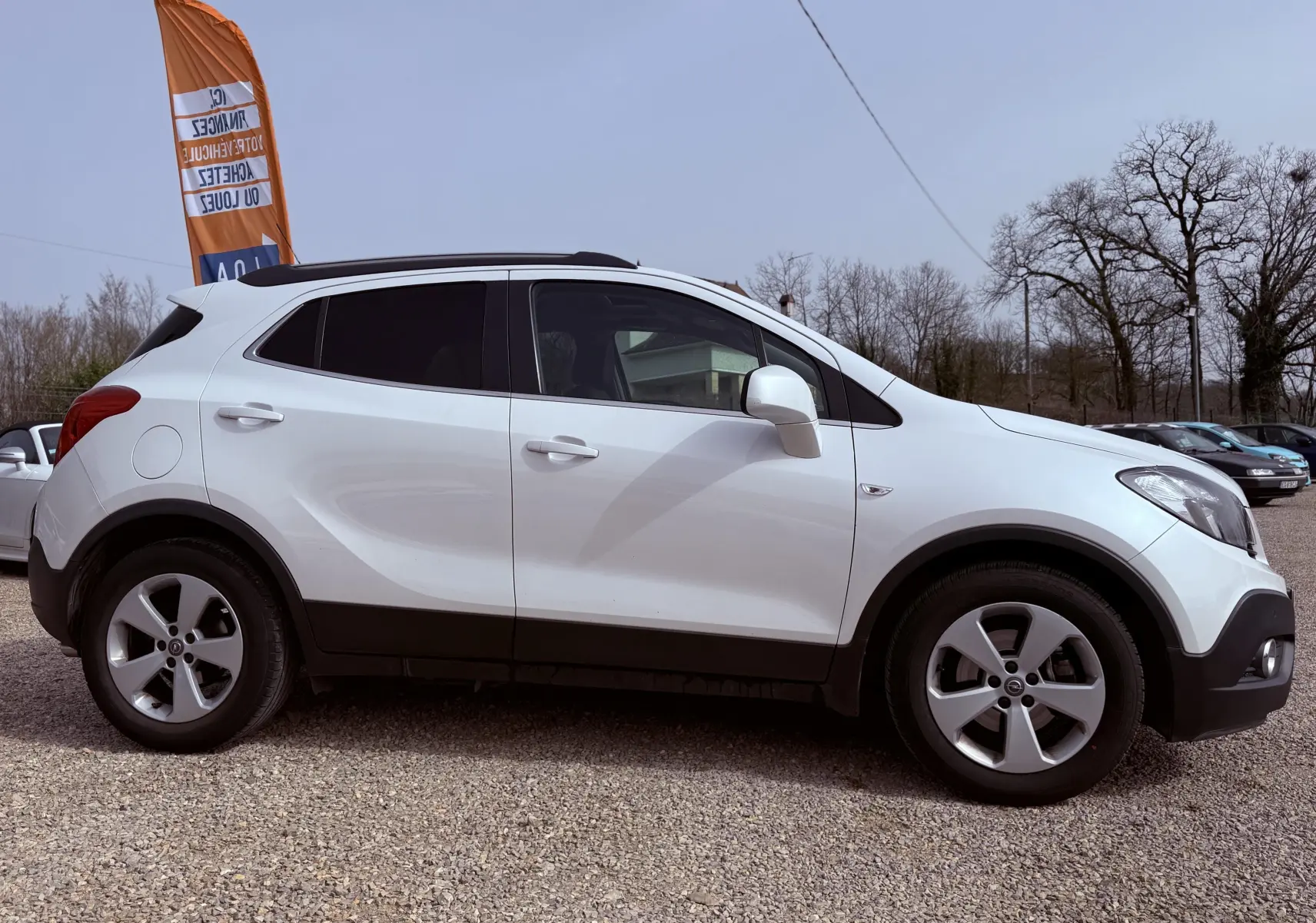 Vue latérale droite d’un Opel Mokka X blanc 5 portes garé sur un sol gravillonné avec un drapeau orange en arrière-plan.