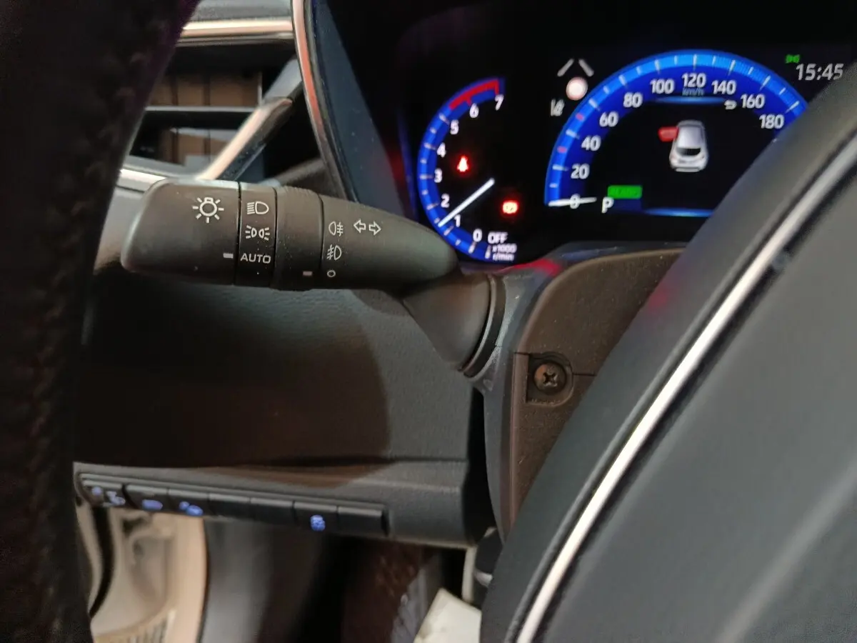 Gros plan sur le combiné d'instruments numérique bleu et le commodo d'éclairage à gauche du volant dans une Toyota Corolla hybride blanche.