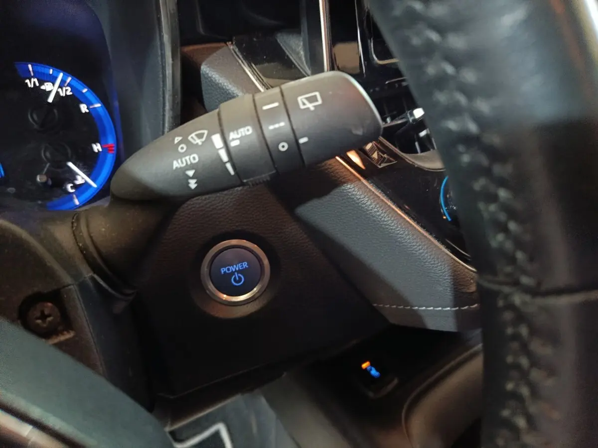 Gros plan intérieur sur le bouton start/stop bleu et la commande d’essuie-glace à gauche du volant cuir d’une Toyota Corolla hybride.