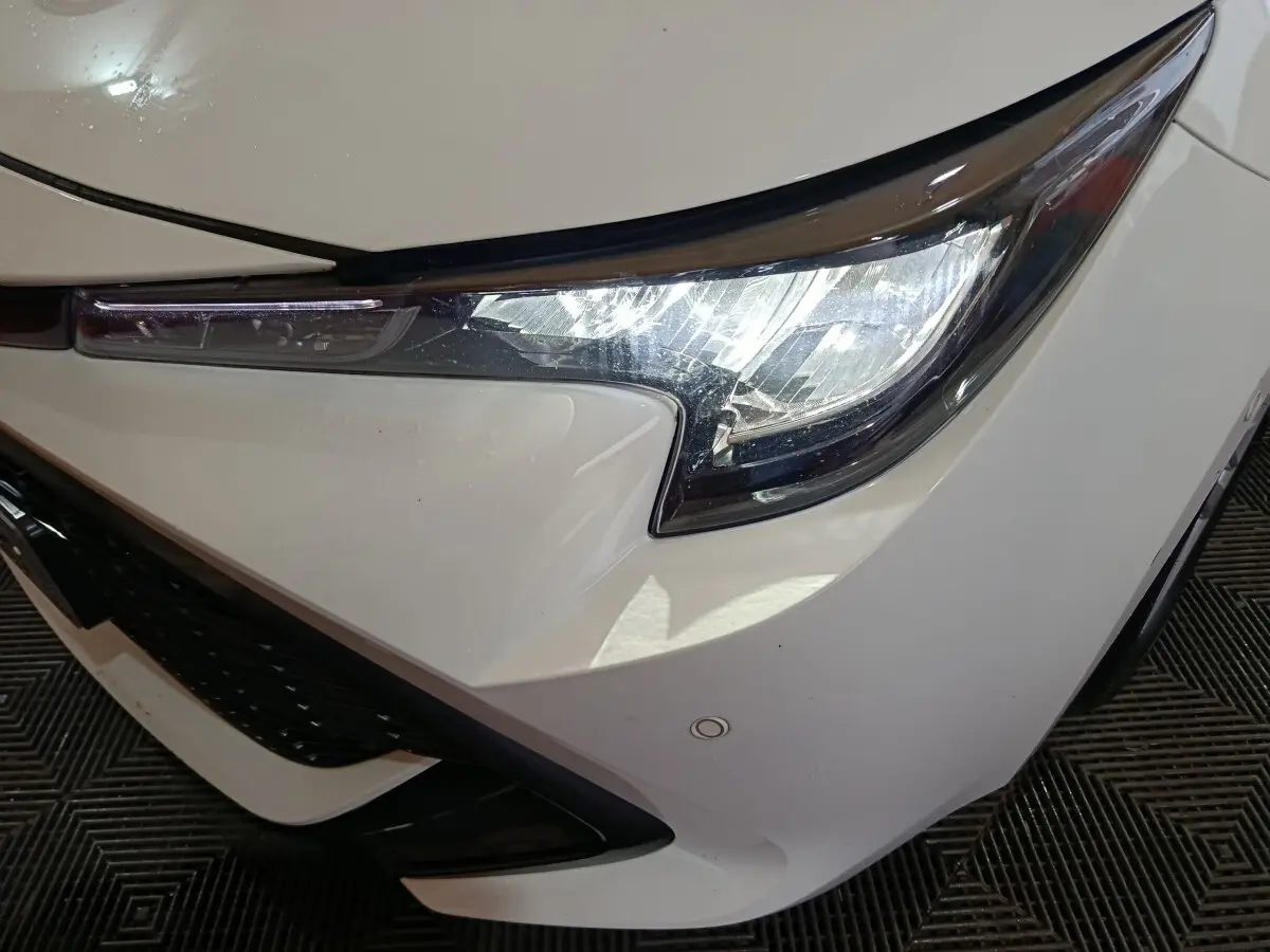 Gros plan sur le phare avant droit blanc d'une Toyota Corolla hybride 2022 avec calandre noire et capteur de stationnement visible.