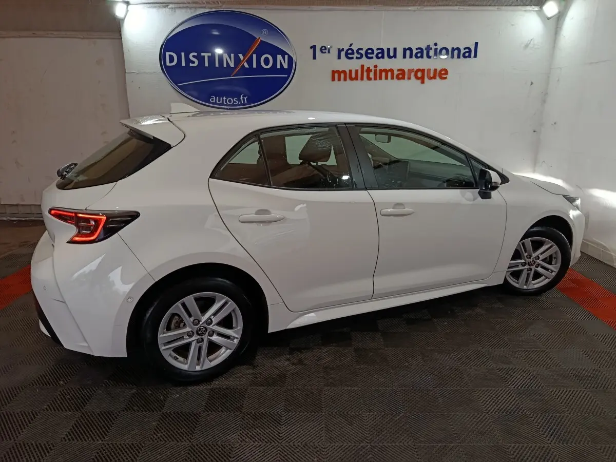 Toyota Corolla hybride blanche vue de profil droit en intérieur avec feux arrière LED allumés et jantes alliage.