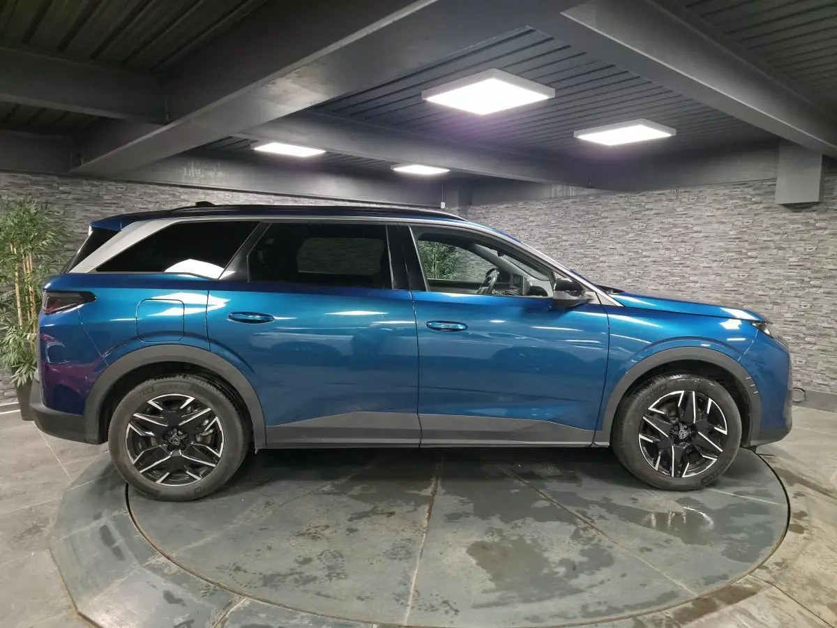 Vue de profil côté gauche d'un Peugeot 5008 bleu métallisé 2025 avec jantes alliage noires et barres de toit.
