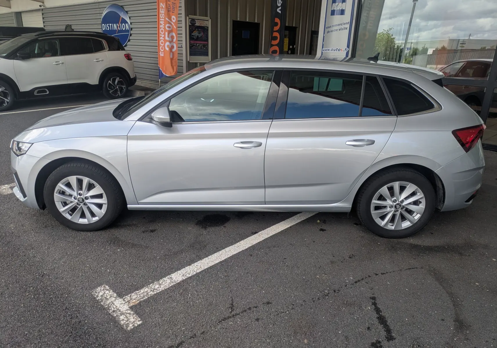 Vue de profil côté gauche d'une Skoda Scala gris reflex garée devant un bâtiment commercial avec vitres teintées.