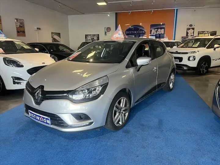 Renault Clio IV gris argent vue 3/4 avant droit dans un showroom avec d'autres véhicules en arrière-plan.