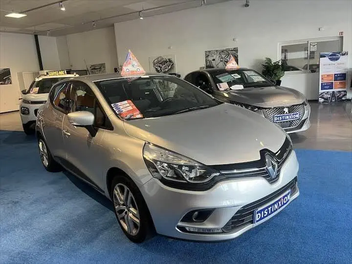 Renault Clio IV gris argent en 3/4 avant droit dans un showroom avec panneau Distinxion visible.