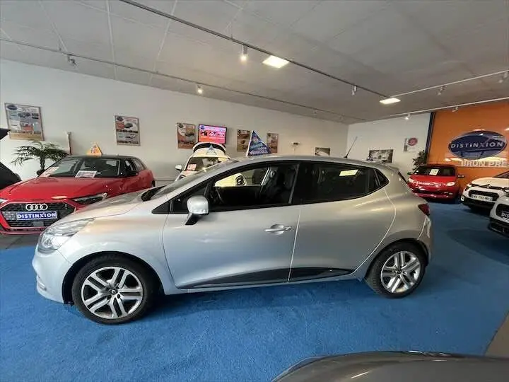 Profil côté gauche d'une Renault Clio IV gris argent exposée en showroom sur sol bleu, entourée d'autres véhicules.