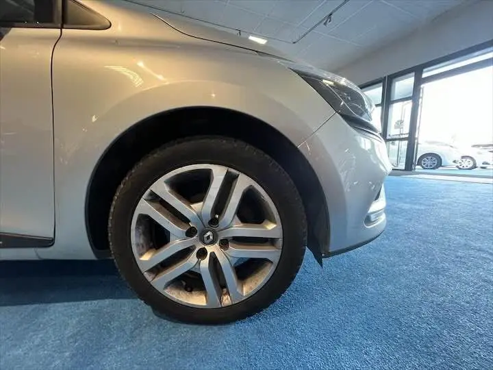 Gros plan sur la roue avant gauche d'une Renault Clio IV gris argent, avec partie avant visible en intérieur showroom.